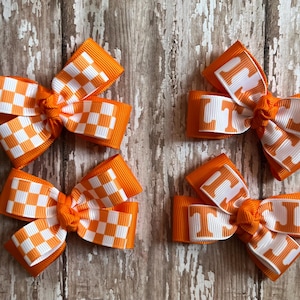 May include: Four orange and white hair bows. Two bows have a checkered pattern, and two have the letters "UT" in white. The bows are made of ribbon and are tied in the center.