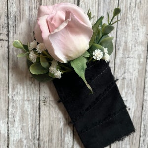 May include: A single pink rose with white baby's breath and green foliage is attached to a black fabric with frayed edges.