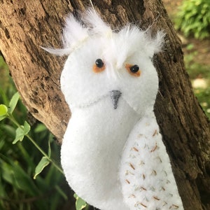 May include: A white felt owl ornament with orange eyes and a gray beak, perched on a tree branch. The owl has feathery tufts and glitter accents on its wings, creating a whimsical, handcrafted look. A decorative item.
