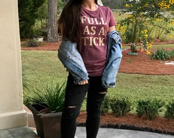 Puede incluir: Una mujer con una camiseta granate con el texto "FULL AS A TICK" en letras doradas, una chaqueta vaquera, unos vaqueros negros rotos y mocasines marrones.
