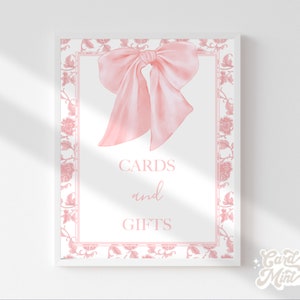 May include: A white sign with a pink floral border and a pink bow. The sign says "Cards and Gifts".