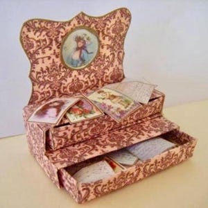 Peut inclure: Une boîte décorative à trois niveaux avec un motif floral rose et brun. Le niveau supérieur a un ovale découpé avec une image vintage d'une fille. La boîte est ouverte et remplie de cartes postales anciennes.