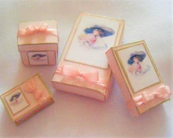 May include: Four pink boxes with gold trim and ribbon. The boxes have images of women in hats and dresses. The boxes are arranged in a row on a white surface.