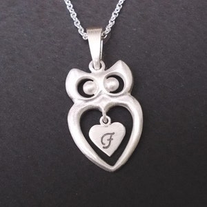 Puede incluir: Un collar con colgante de búho plateado. El búho tiene un cuerpo en forma de corazón con un colgante de corazón más pequeño en el interior, grabado con la letra "F". La cadena del collar también es plateada, sobre un fondo negro.