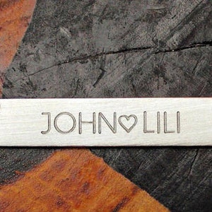 May include: Silver necklace with a rectangular bar pendant engraved with the names "JOHN" and "LILI" separated by a heart symbol.