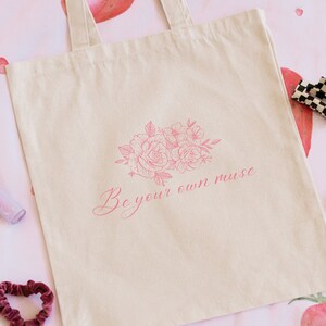 Op de afbeelding: Een beige canvas tote bag met een roze bloemenontwerp en de tekst "Be your own muse".