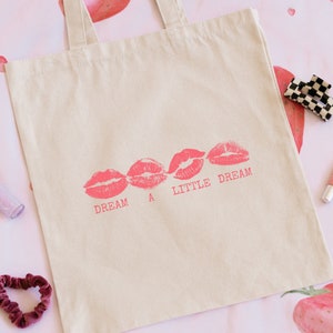Pode incluir: Uma bolsa tote de lona branca com um design de estampa de l&aacute;bios rosa e o texto "DREAM A LITTLE DREAM".