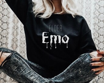 May include: A black sweatshirt with the text "ELDER Emo" printed in white with dripping paint accents. The sweatshirt is being worn by a person wearing ripped jeans and leopard print sneakers.