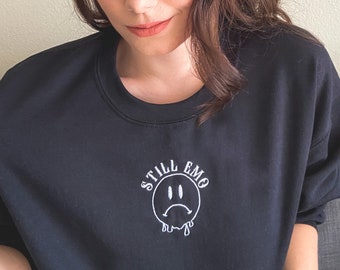 May include: A black sweatshirt with a white embroidered design of a sad face and the text "Still Emo".