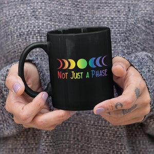 Op de afbeelding: Zwarte koffiemok met een regenboog maanfasen ontwerp en de tekst "Not Just A Phase".