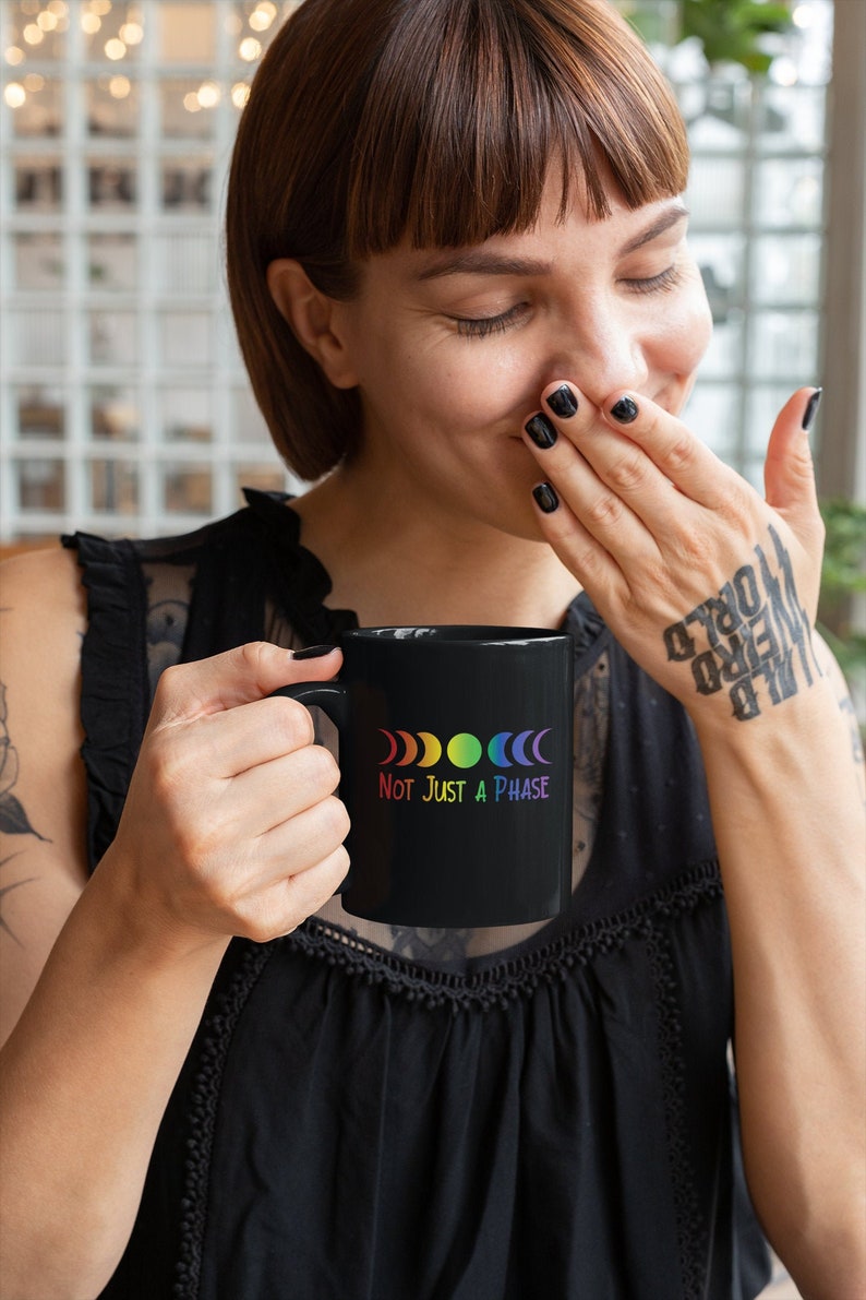 Op de afbeelding: Een zwarte koffiemok met een regenboog maanfasen ontwerp en de tekst "Not Just A Phase".