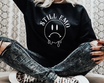 May include: Black sweatshirt with a white graphic of a sad face and the text "STILL EMO". The model is wearing the sweatshirt with ripped jeans and leopard print sneakers.