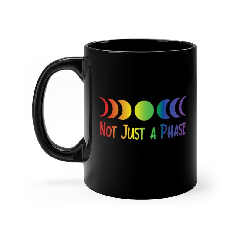 Op de afbeelding: Zwarte koffiemok met een regenboog maanfasen ontwerp en de tekst "NOT JUST A PHASE".