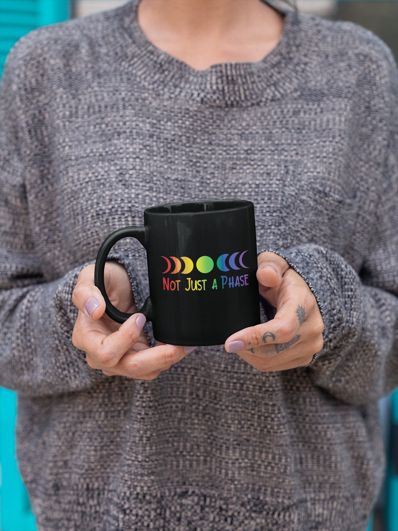 Op de afbeelding: Zwarte koffiemok met een regenboog maanfasen ontwerp en de tekst "Not Just A Phase".