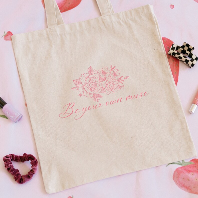 Op de afbeelding: Een beige canvas tote bag met een roze bloemenontwerp en de tekst "Be your own muse".