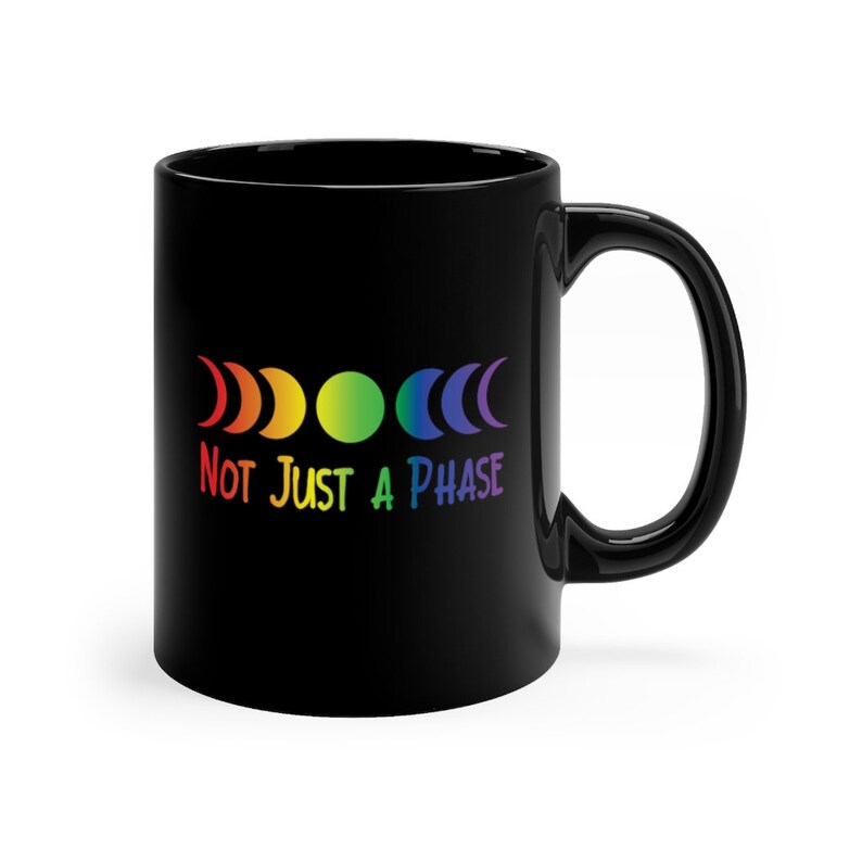 Op de afbeelding: Zwarte keramische koffiemok met een regenboog maanfasen ontwerp en de tekst "Not Just A Phase".