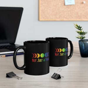 Op de afbeelding: Twee zwarte koffiemokken met een regenboog maanfasen ontwerp en de tekst "Not Just A Phase".
