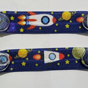 Pode incluir: Dois suportes de máscara de tecido azul com um padrão espacial com foguetes, planetas e estrelas. Cada suporte tem clipes de plástico preto em ambas as extremidades para prender a uma máscara.