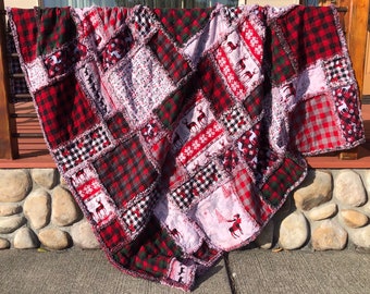 May include: A red and black plaid, white and pink, and white and gray patchwork quilt with a Christmas reindeer pattern. The quilt is folded and draped over a stone wall.