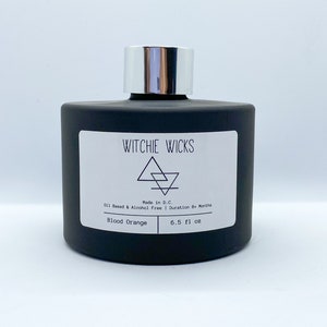 May include: A black glass bottle with a silver lid and a white label that reads "WITCHIE WICKS" with a geometric symbol and the text "Made in D.C. Oil Based & Alcohol Free | Duration 6+ Months Blood Orange 6.5 fl oz".