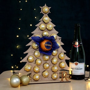 May include: A cardboard Christmas tree advent calendar with 24 numbered doors, each containing a chocolate. The tree is decorated with a blue bow and a large orange chocolate. A bottle of champagne is to the right of the tree.