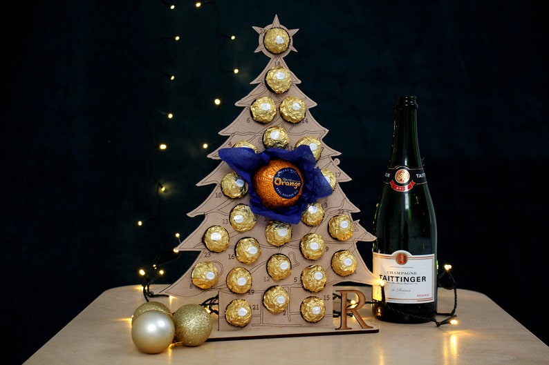 May include: A cardboard Christmas tree advent calendar with 24 numbered doors, each containing a chocolate. The tree is decorated with a blue bow and a large orange chocolate. A bottle of champagne is to the right of the tree.
