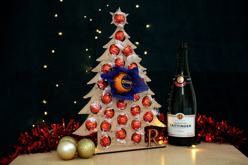 May include: A wooden Christmas tree advent calendar with 24 numbered doors, filled with red and orange chocolate truffles. A bottle of Taittinger Champagne is in the foreground, with red and gold tinsel.
