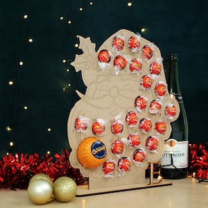 May include: A wooden Santa Claus advent calendar with 25 numbered compartments filled with chocolate candies. The calendar is standing on a table with a bottle of champagne and red tinsel.
