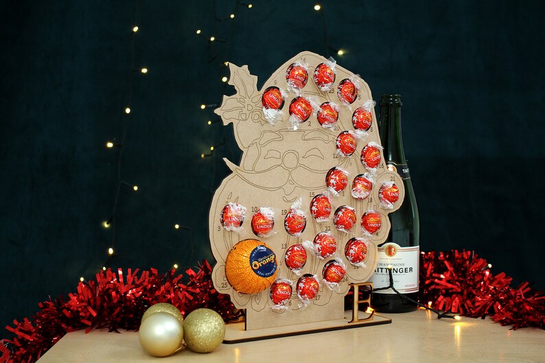 May include: A wooden Santa Claus advent calendar with 25 numbered compartments filled with chocolate candies. The calendar is standing on a table with a bottle of champagne and red tinsel.