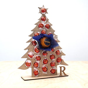 May include: A wooden Christmas tree advent calendar with 24 numbered compartments, each filled with a chocolate. The tree is decorated with a blue bow and a star on top. The calendar is personalized with the letter "R".