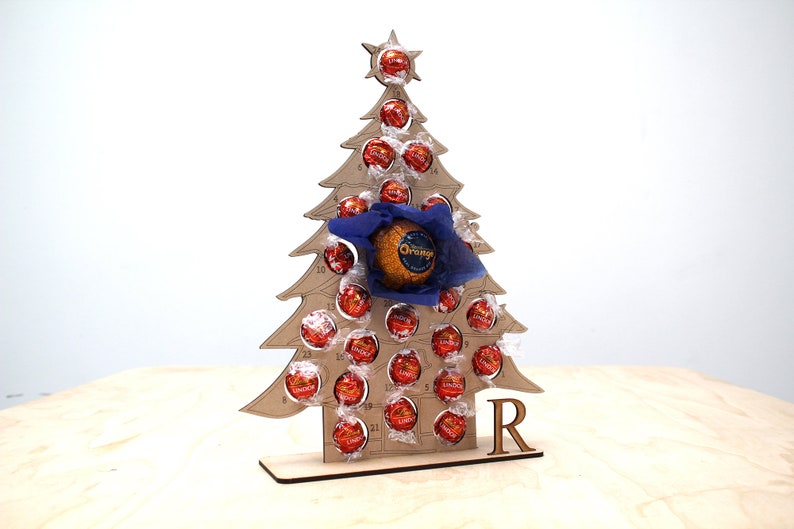 May include: A wooden Christmas tree advent calendar with 24 numbered compartments, each filled with a chocolate. The tree is decorated with a blue bow and a star on top. The calendar is personalized with the letter "R".