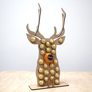 May include: A wooden reindeer shaped advent calendar with 24 numbered compartments filled with gold foil wrapped chocolates. The reindeer is standing on a wooden base with the number "11" printed on it.