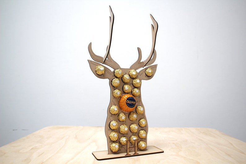 May include: A wooden reindeer shaped advent calendar with 24 numbered compartments filled with gold foil wrapped chocolates. The reindeer is standing on a wooden base with the number "11" printed on it.