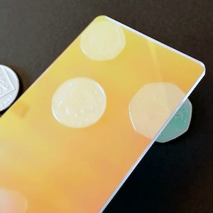 Puede incluir: Una tarjeta de plástico transparente con un acabado holográfico. La tarjeta tiene tres formas circulares que parecen ser monedas, pero en realidad son parte del diseño. La tarjeta es de color amarillo-naranja con un efecto degradado.