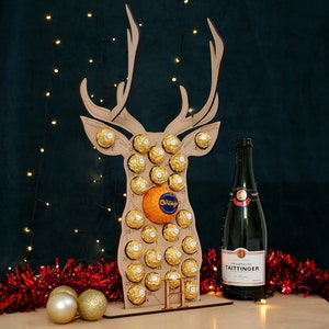 May include: A wooden reindeer advent calendar with 24 chocolate balls and an orange in the center. The calendar is decorated with gold glitter and has a bottle of Taittinger champagne in front of it.