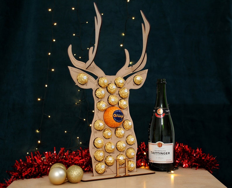May include: A wooden reindeer advent calendar with 24 chocolate balls and an orange in the center. The calendar is decorated with gold glitter and has a bottle of Taittinger champagne in front of it.