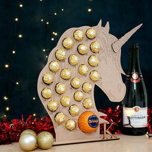May include: A wooden unicorn-shaped advent calendar with 24 numbered compartments filled with chocolate balls. An orange is in the bottom compartment. A bottle of Taittinger champagne is to the right of the calendar. Red and gold tinsel is in the foreground.