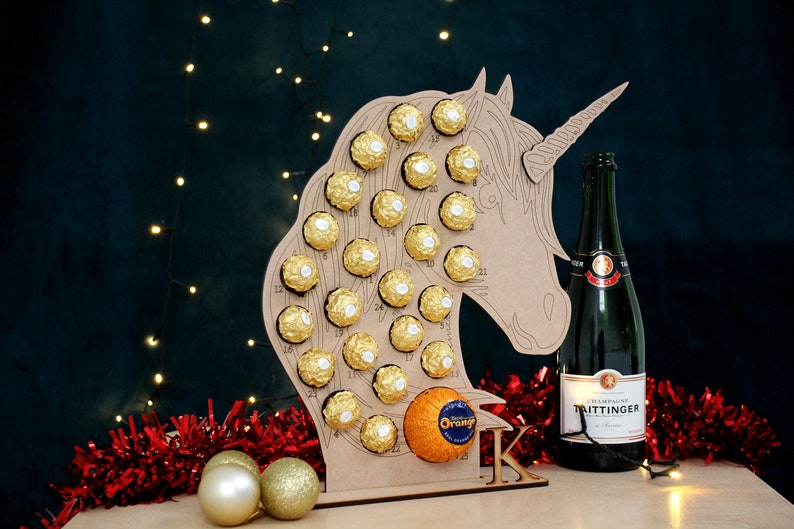 May include: A wooden unicorn-shaped advent calendar with 24 numbered compartments filled with chocolate balls. An orange is in the bottom compartment. A bottle of Taittinger champagne is to the right of the calendar. Red and gold tinsel is in the foreground.