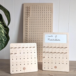May include: Three wooden meditation calendars with pegs and a larger wooden board. One calendar has the text "1 month of Meditation". The calendars are light brown and have numbers 1-31. The larger board has the months of the year.