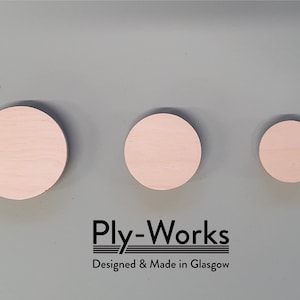Puede incluir: Tres círculos de madera rosa claro de diferentes tamaños sobre un fondo gris. El texto "Ply-Works Designed & Made in Glasgow" está impreso debajo.