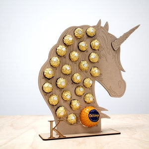 May include: A wooden unicorn-shaped advent calendar with 25 numbered compartments filled with gold-wrapped chocolates. The calendar is standing on a wooden base with the letter "K" printed on it.