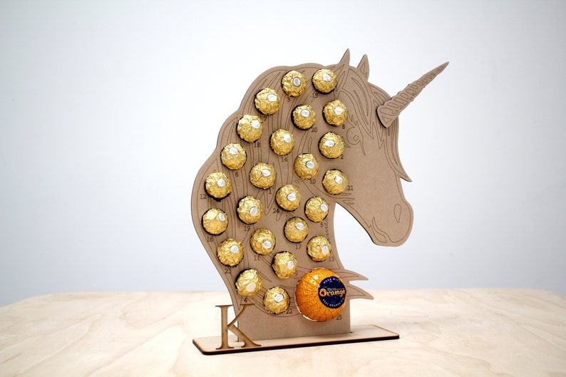 May include: A wooden unicorn-shaped advent calendar with 25 numbered compartments filled with gold-wrapped chocolates. The calendar is standing on a wooden base with the letter "K" printed on it.