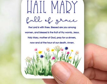 May include: A white card with a blue border and the text "HAIL MARY full of grace" in blue lettering. The card has a watercolor illustration of green grass and colorful flowers.