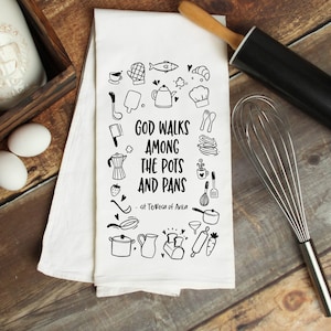 May include: A white kitchen towel with black ink illustrations of kitchen utensils and food. The text reads "God walks among the pots and pans - St. Teresa of Avila".