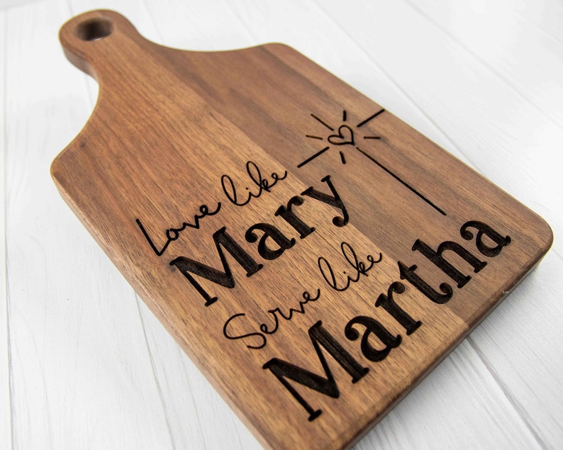 May include: A wooden cutting board with the text "Love like Mary, Serve like Martha" engraved on it. The board has a handle and a heart with a starburst design.