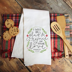 May include: White kitchen towel with the text "I will be glad & rejoice in your love" surrounded by a green wreath. The towel is next to wooden cooking utensils and chocolate chip cookies on a plaid cloth.