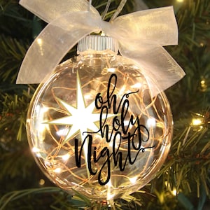 May include: A clear glass Christmas ornament with a white star and the words "Oh Holy Night" written in black calligraphy. The ornament is decorated with warm white fairy lights.