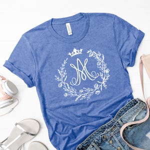 May include: A blue t-shirt with a white floral wreath design and a crown above a stylized letter "M". The shirt is made of a soft material and has rolled-up sleeves. The shirt is paired with denim jeans and accessories.