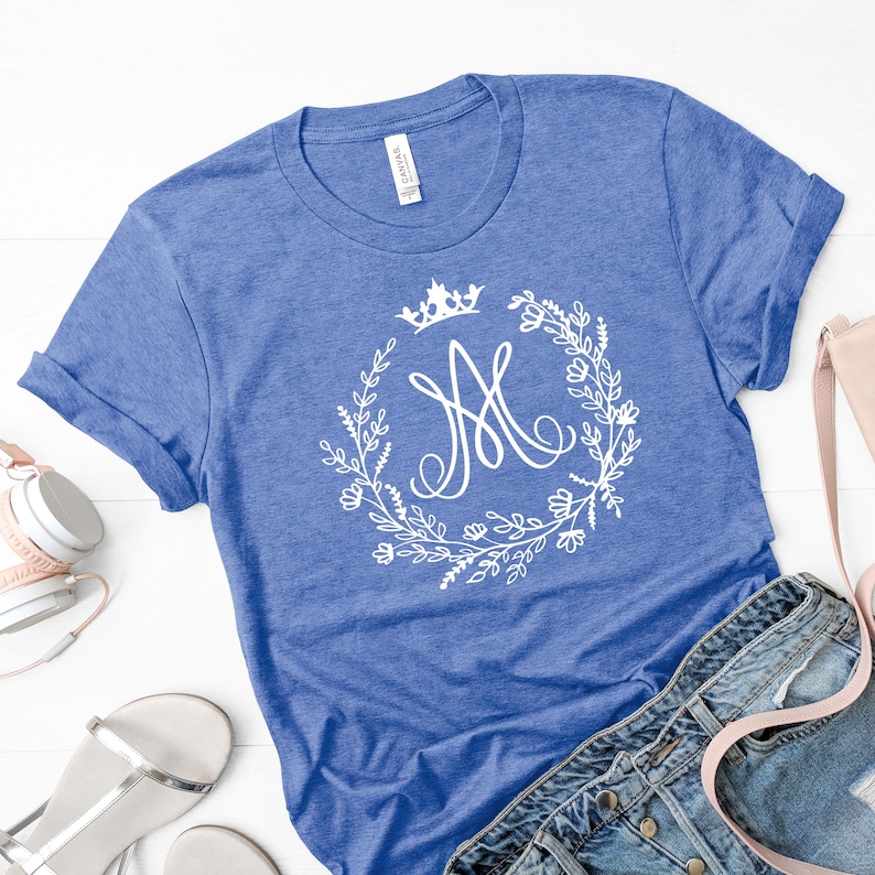 May include: A blue t-shirt with a white floral wreath design and a crown above a stylized letter "M". The shirt is made of a soft material and has rolled-up sleeves. The shirt is paired with denim jeans and accessories.