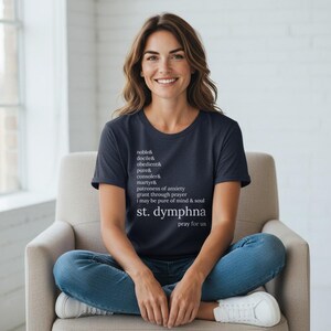 May include: A navy blue t-shirt with white text. The text includes the words "noble", "docile", "obedient", "pure", "consoler", "martyr", "patroness of anxiety", "st. dymphna", and "pray for us". The person in the image is wearing blue jeans and white trainers.